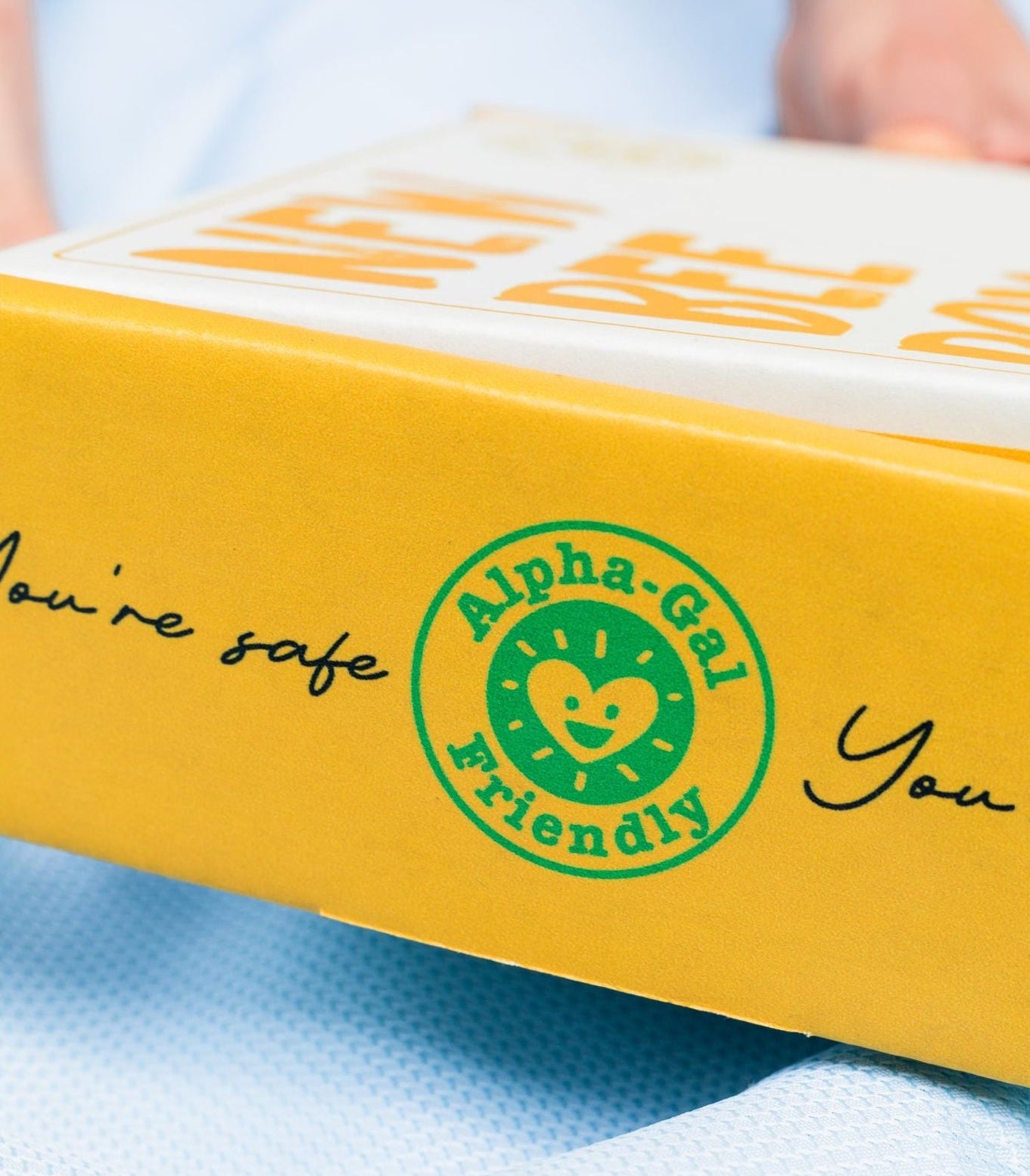 A close-up of a yellow product box with a green circular 'Alpha-Gai Friendly' logo and partial black script text, resting on a light blue textured surface.