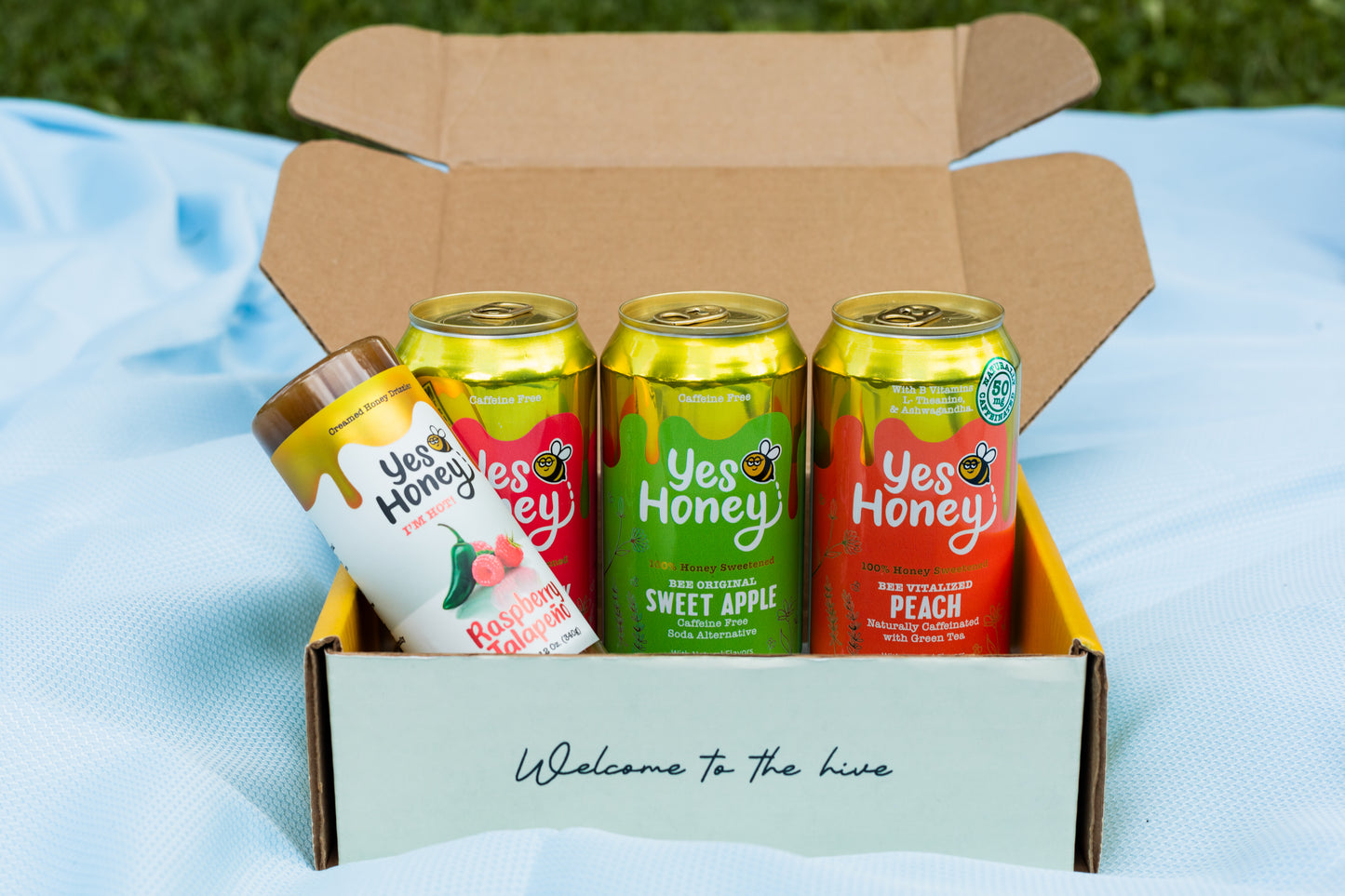 A cardboard gift box on a light blue blanket outdoors, containing three Yes Honey canned honey sodas: a white bottle with a gold lid labeled Raspberry Jalapeño hot honey, a green can labeled Sweet Apple, an orange can labeled Peach, and a red  can with labeled Strawberry.