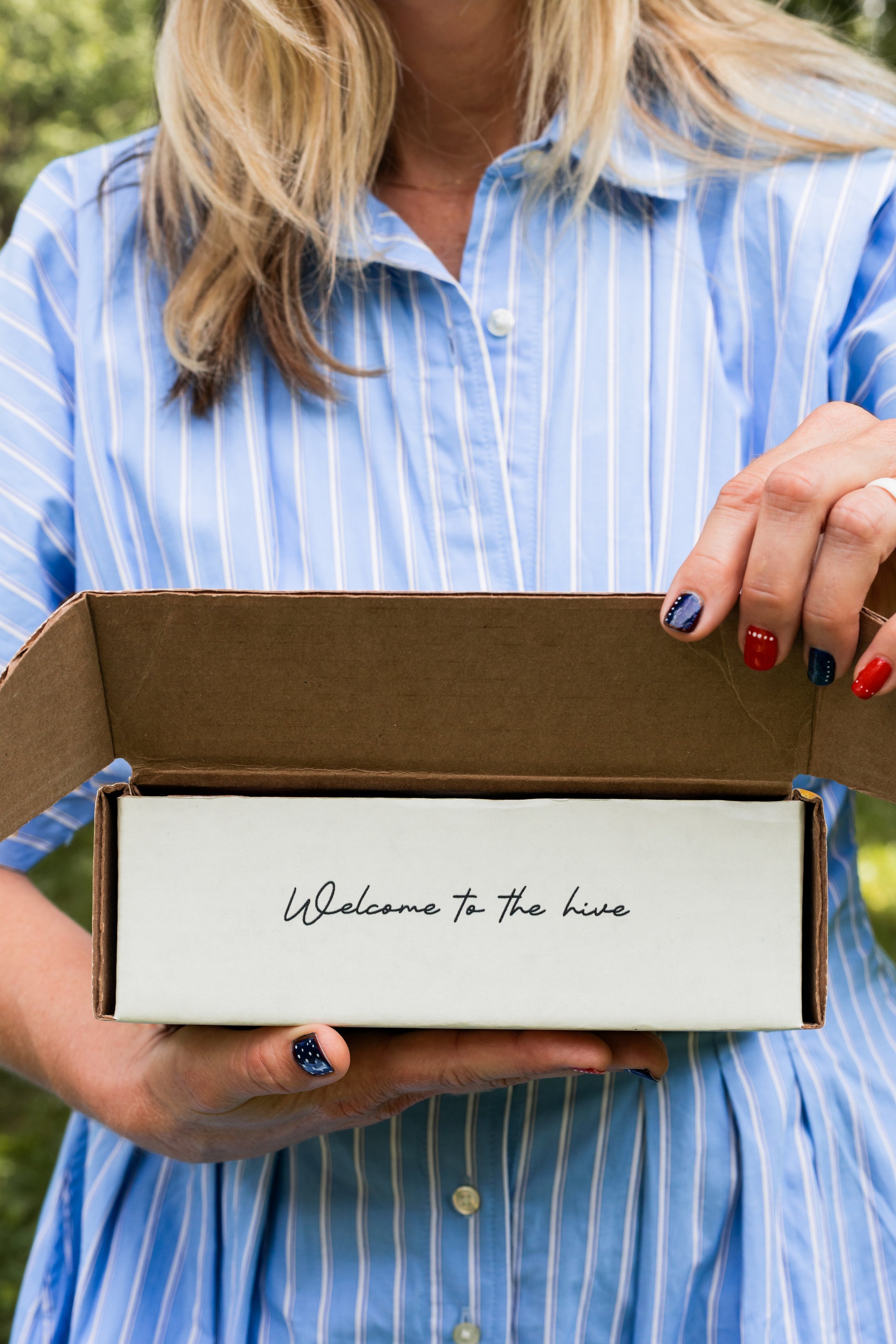 A woman in a blue and white striped shirt holds open a white  cardboard box containing the handwritten text 'Welcome to the hive'.
