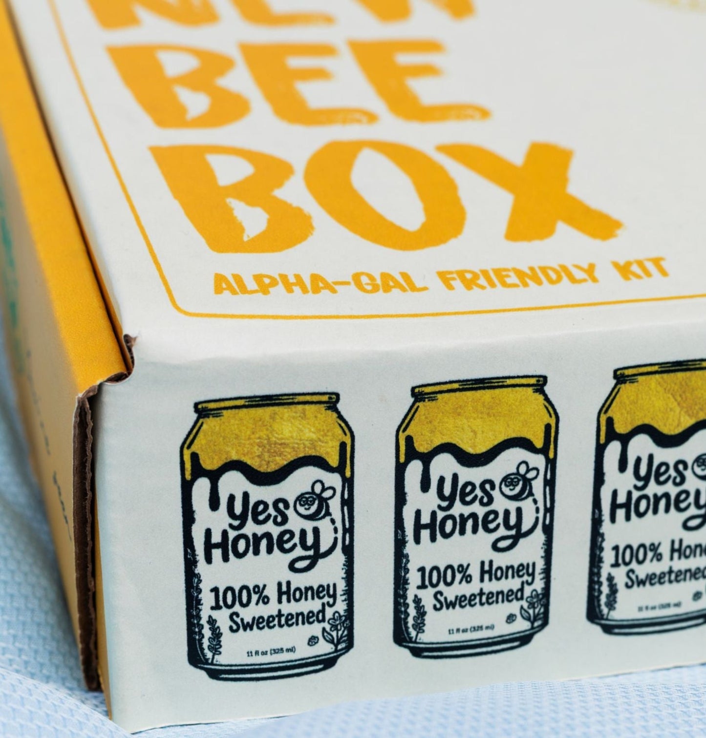 A close-up of a 'New Bee Box' kit package, showing the side panel with illustrations of 'Yes Honey' honey cans and the top corner displaying 'BEE BOX' in bold yellow text.