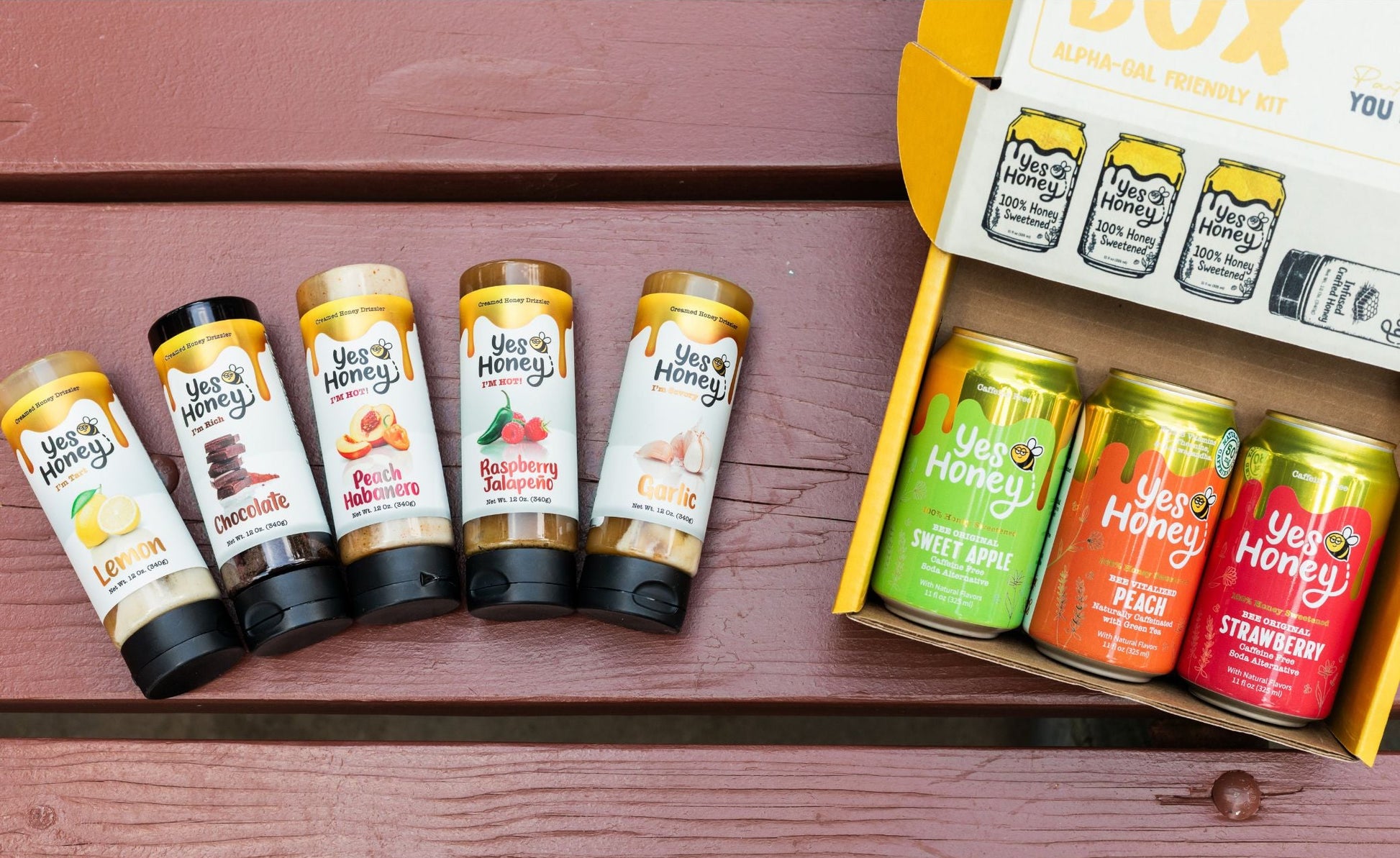 A boxed honey flavor assortment set displayed on a reddish-brown wooden surface, with four small bottles lined up on the left and an open box containing three colorful cans on the right. The labels read 'Yes Honey' and show flavors including lemon, chocolate, apple, peach, and strawberry.