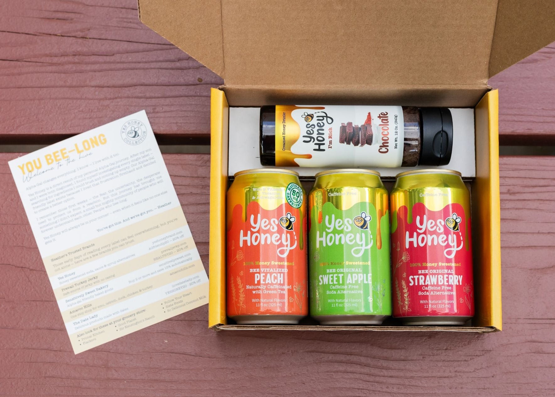 A Yes Honey beverage gift box shown from above on a reddish-brown wooden surface, with the box open to reveal three colorful cans of honey sweetened soda - a can of apple, a can of strawberry, and a can of peach, a white bottle of chocolate honey, and a printed insert card.